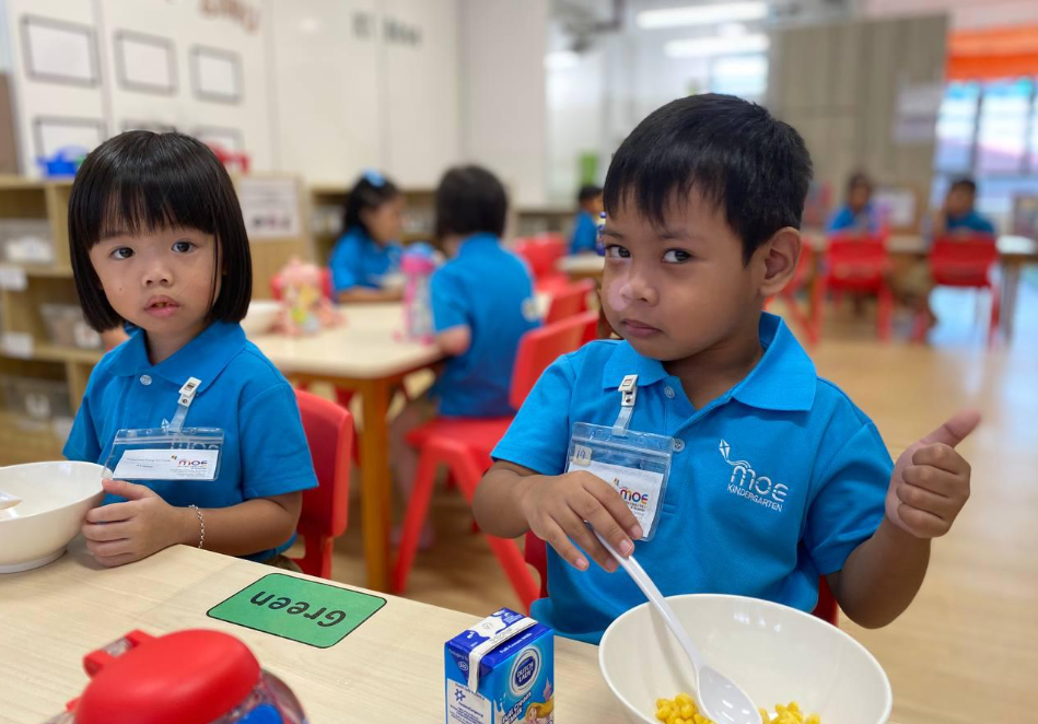 Two children in blue uniforms sit at a classroom table with bowls and food; boy gives a thumbs-up.